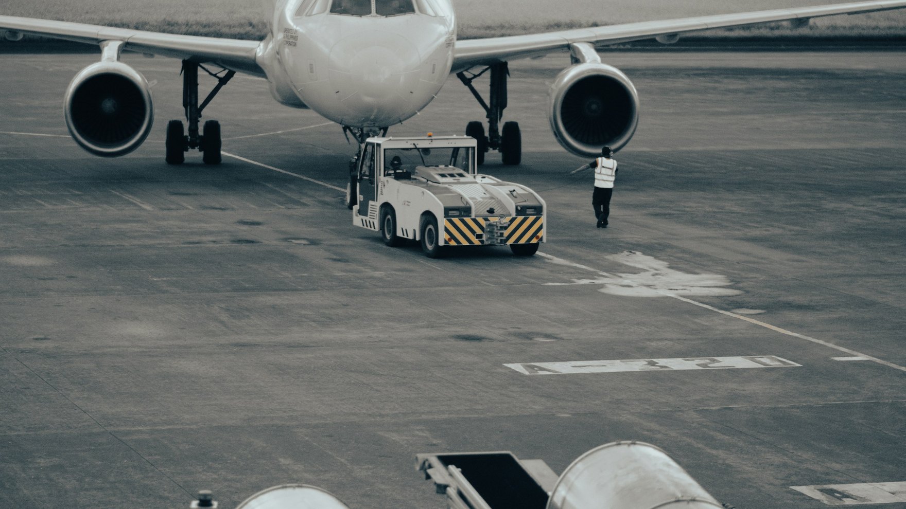 A tow tug pushes back a white passenger jet while a ground crew member walks beneath the engine.